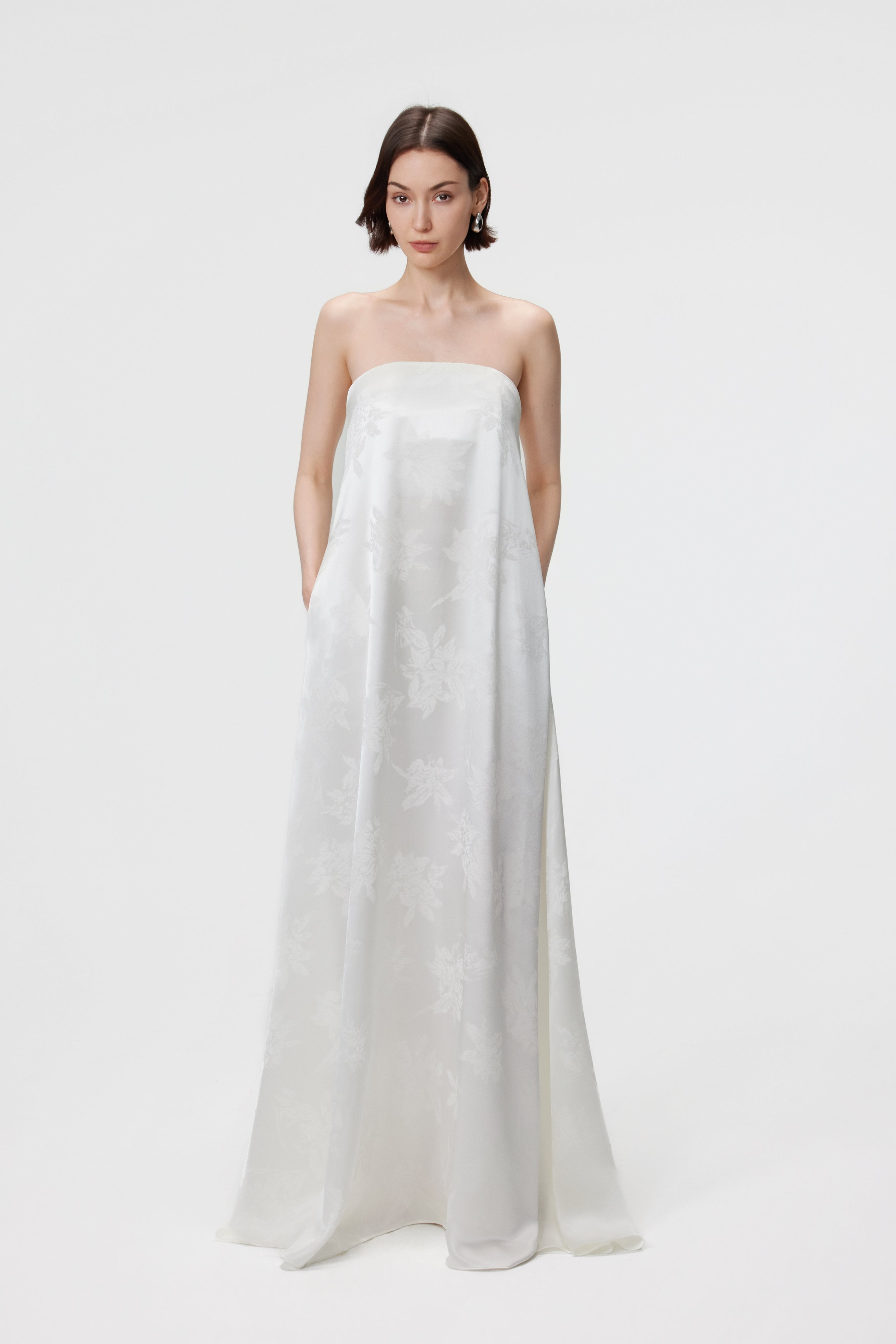Full-length front view of a model wearing the Aenso Iris Jacquard Strapless Maxi Dress in White. The image highlights the off-the-shoulder Bardot neckline, structured bustier, and the luminous tonal floral pattern of the premium Naia™ Acetate fabric.