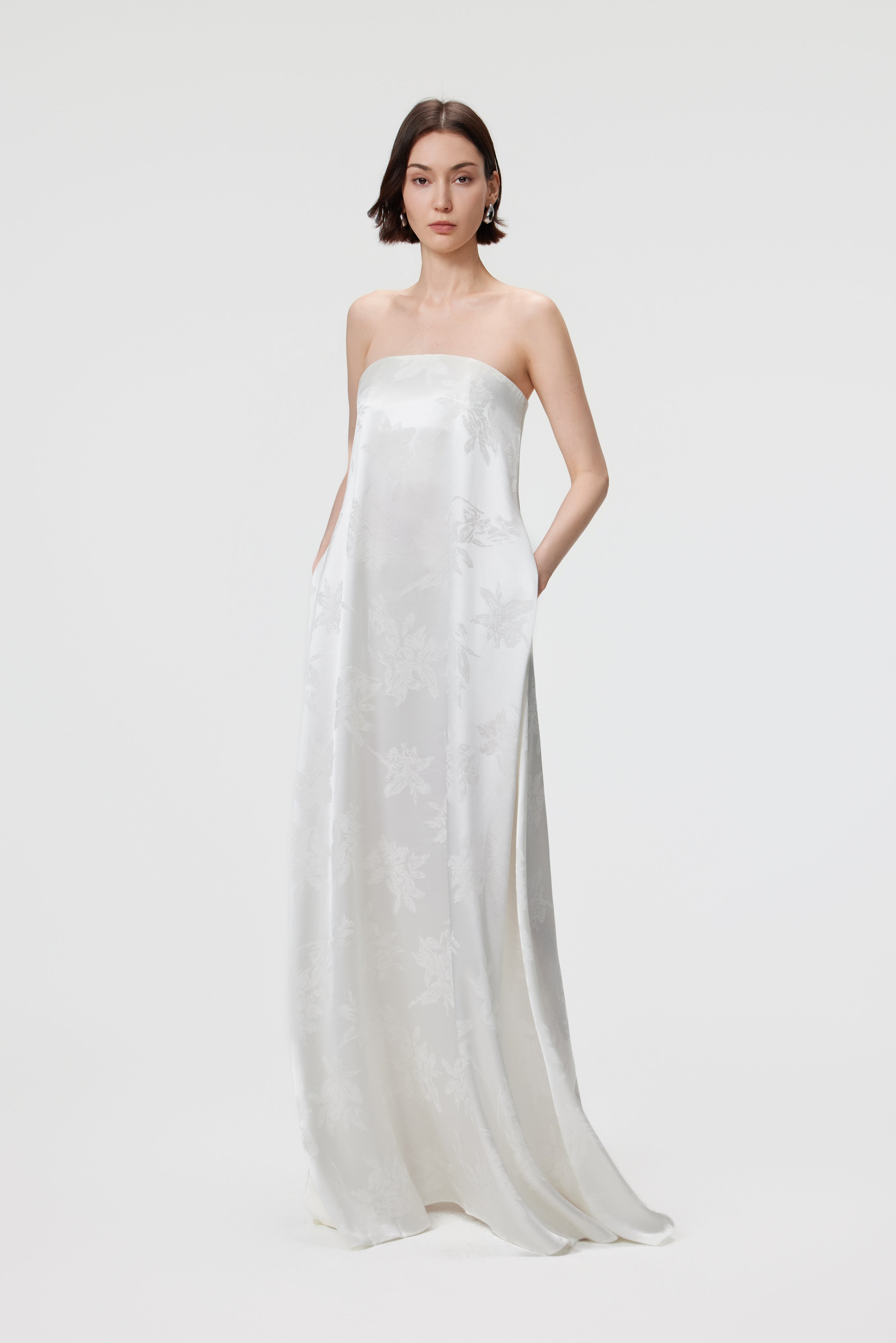 A full-length view of the Iris Jacquard Strapless Maxi Dress in White, featuring a model with hands in the functional internal side pockets. The shot highlights the relaxed, straight-cut silhouette and the effortless movement of the floral jacquard fabric, demonstrating the blend of formal occasion wear with modern utility.