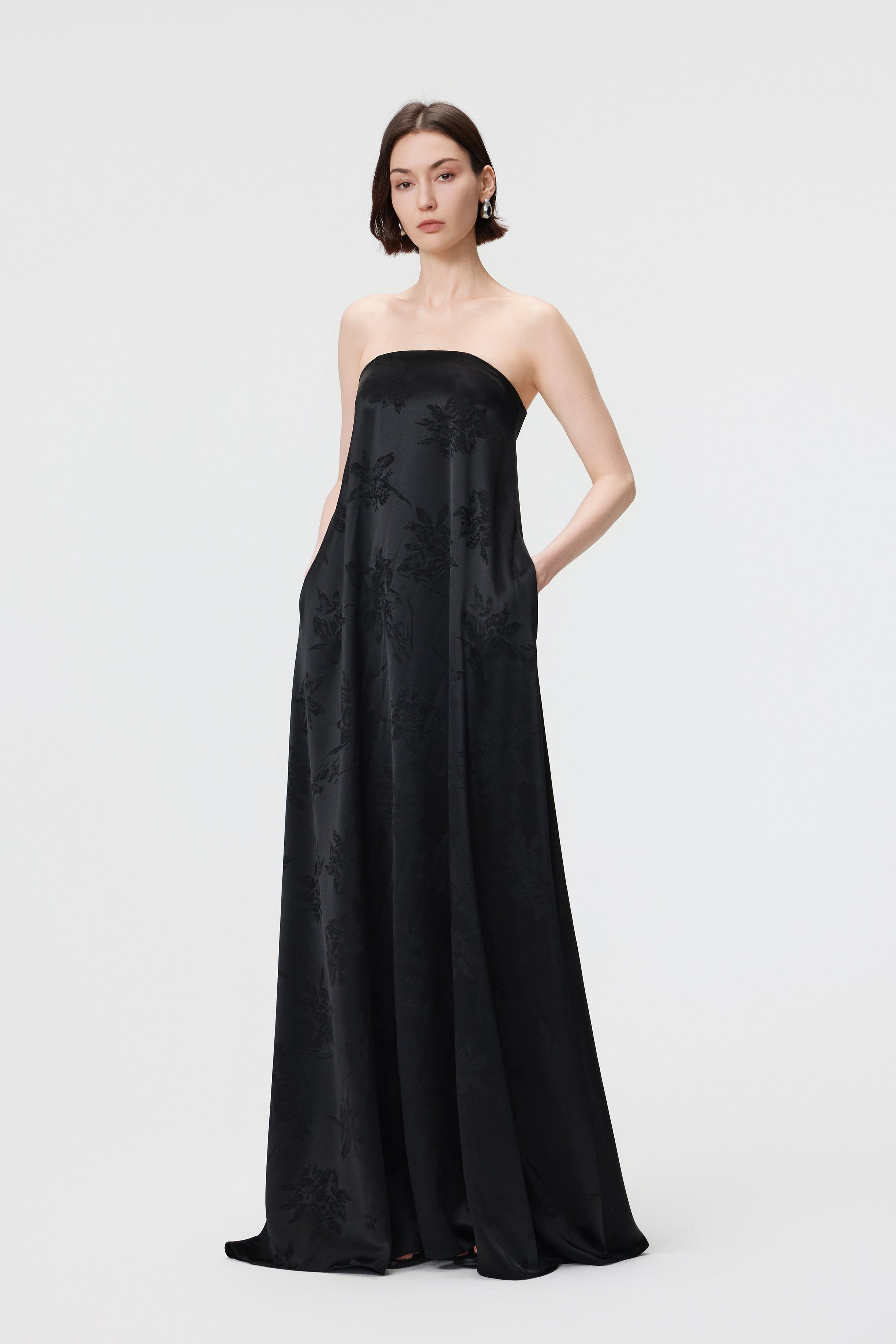 Front view of the AENSO Iris gown in Black; the model stands with hands in the discreet side-seam pockets, showcasing the effortless intersection of functional detail and the sculptural column silhouette in dimensional floral jacquard.