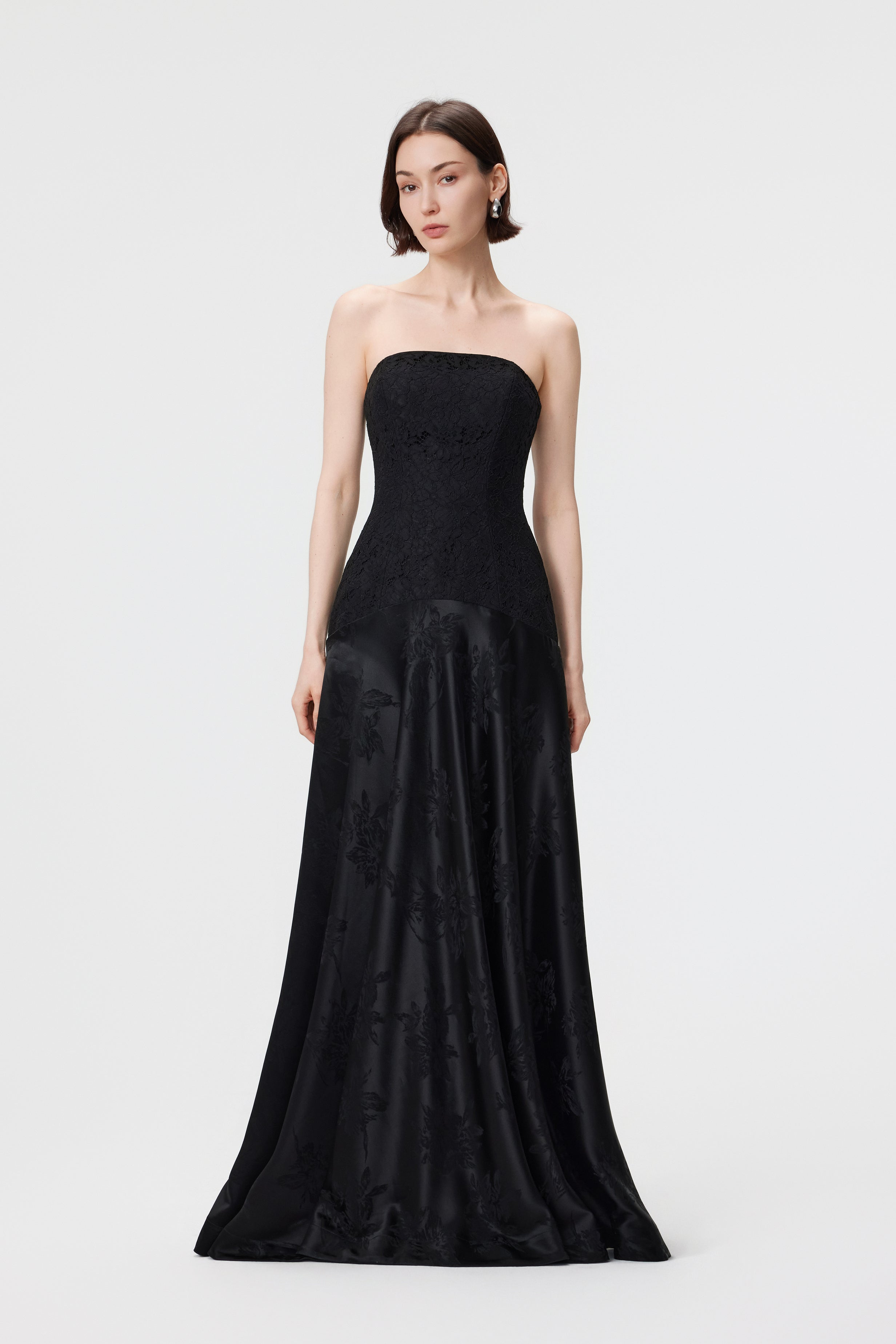 A statuesque front-on view of the Vera gown in Black, showcasing the precision-paneled lace bodice and the weighted, rhythmic drape of the floral jacquard skirt.