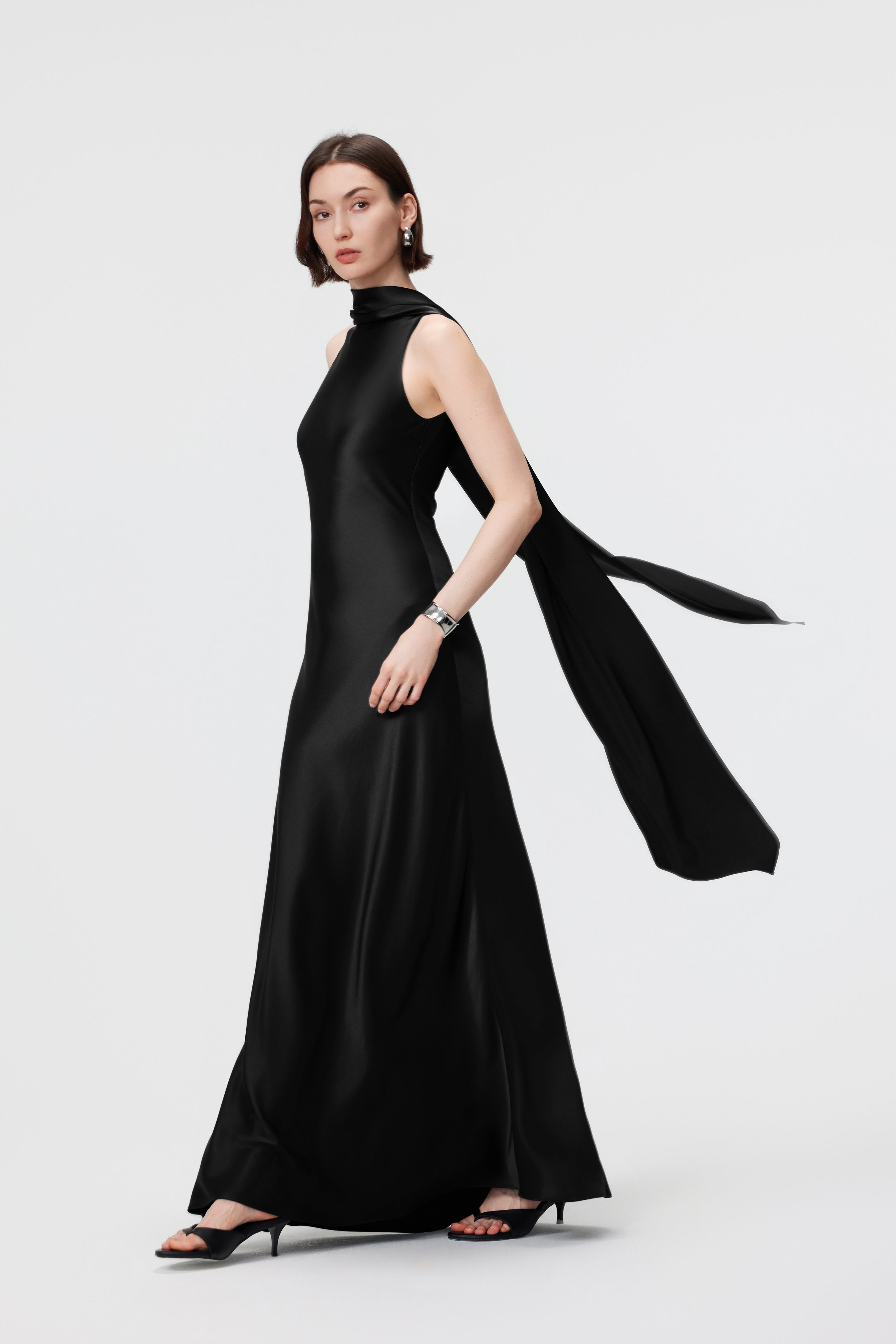 Side profile of the AENSO Elara gown in Black in motion; the bias-cut construction creates a rhythmic, liquid-like trail that highlights the rich saturation of the Naia™ Acetate and the architectural drape of the integrated scarf.