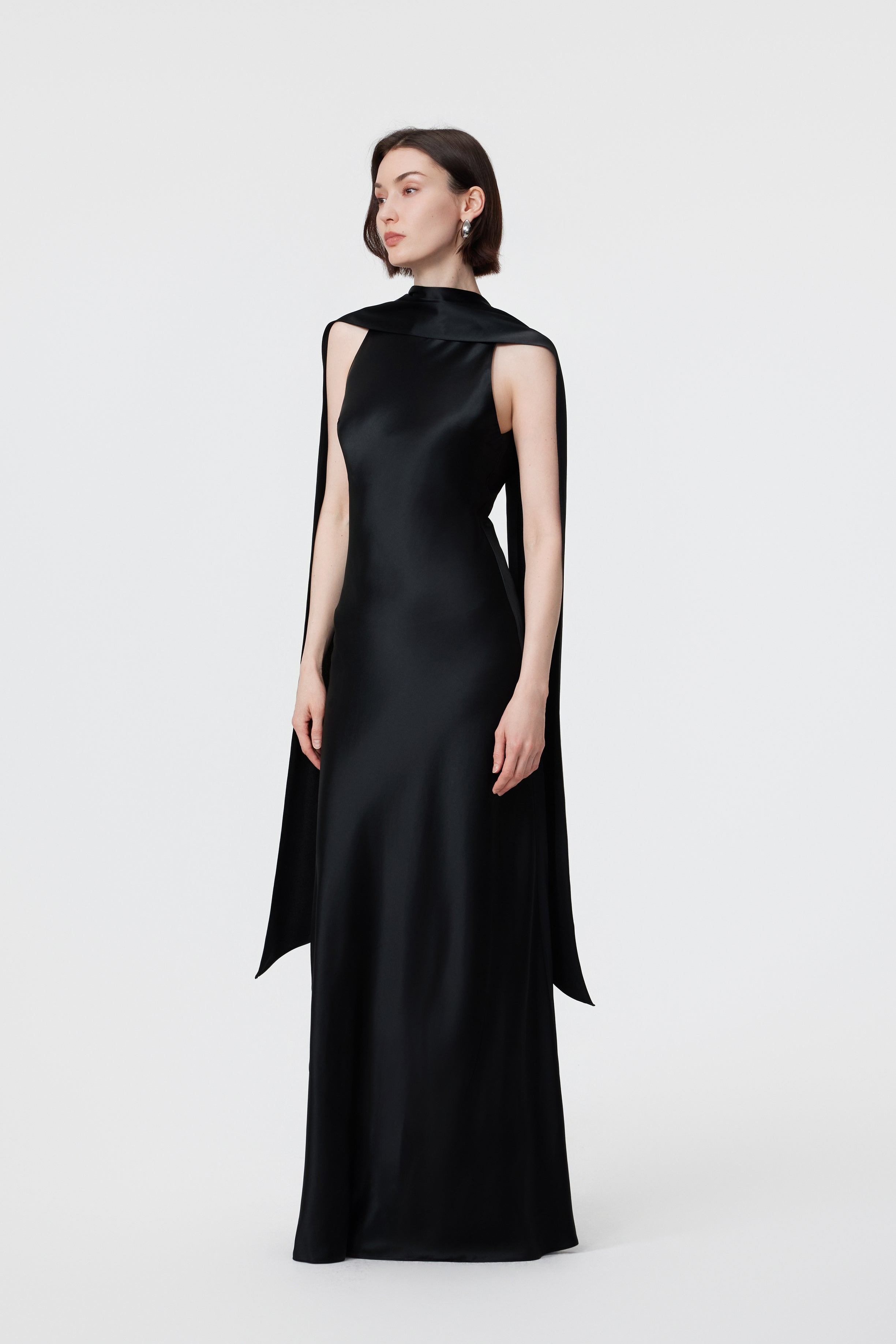 Side profile of the AENSO Elara gown in Black; showcasing the architectural drape of the integrated scarf and the sleek, statuesque lines of the bias-cut silhouette.